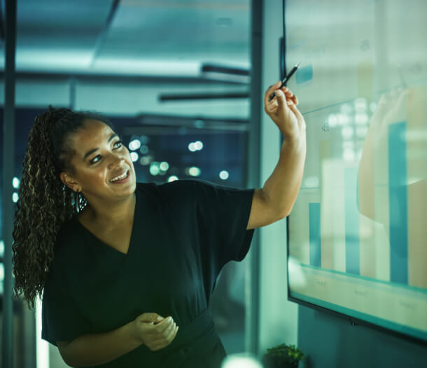 In a dimly lit room, a woman smiles and points at an analytics bar chart on-screen, illustrating Austin Public Relations and Social Media strategy.