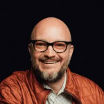 A bearded individual with glasses in a red jacket, representing Austin Public Relations and Social Media Management.