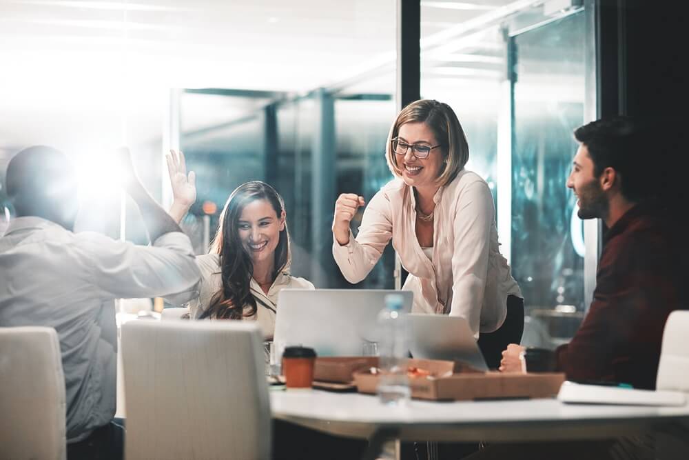 A group of four people in a modern Austin office celebrates around laptops, discussing Public Relations strategies and crisis management.
